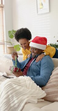 Vertical video: Diverse couple wearing Santa hat unwrapping gift after hugging then talking on sofa