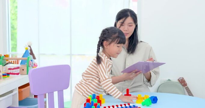 Asian single mother reviews learning result on tablet as preschool daughter continues block activity at table, supporting practice, concentration, sequencing skill, and cognitive growth in play.
