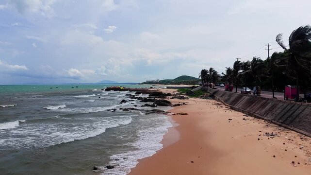 People walk along the shore at Ong Dia Rock Beach. Waves crash on the rocks as the sun shines. The beach is busy with visitors enjoying the warm weather and beautiful scenery.