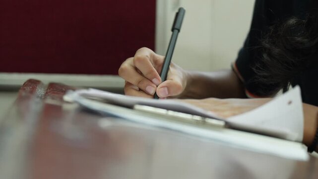 A student writes an examination on paper using a pen at a desk in an Indian classroom. The close-up shot captures the hand holding the pen as it moves across the answer sheet during the test.