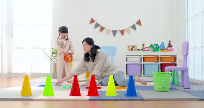 Asian single mother presents colorful ring to preschool daughter beside cone course, encouraging aiming, coordination, playful movement, and stronger child confidence through active learning.