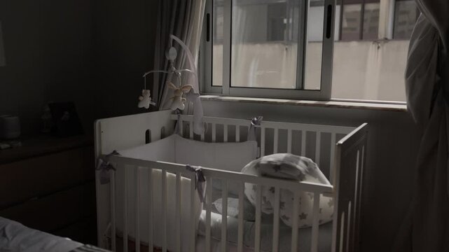 Empty white baby cot with a hanging mobile and cushion beside a curtained bedroom window