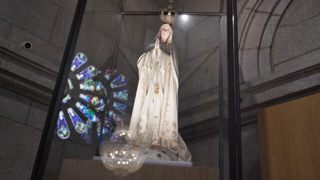 Crowned Virgin Mary statue in a glass display case with stained glass window behind