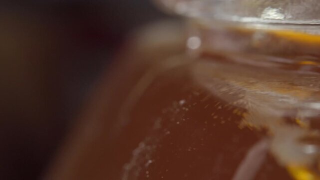 Panning shot moves across glass honey jar filled with golden honey, revealing its rich amber color, glossy texture, reflections, and thick liquid consistency in macro view.