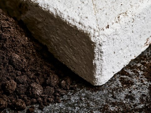 A close-up shot reveals the rough texture of a white brick resting on dark soil, highlighting the contrast between manufactured material and natural earth.