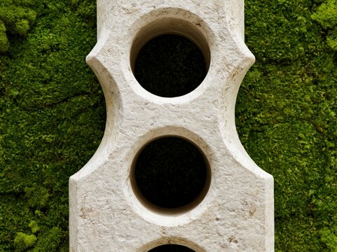 An antique stone architectural element, possibly a fragment of a building or decorative piece, displays three circular openings and textured surfaces, set against a vibrant green moss background.