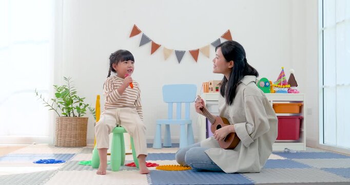 Asian single mother plays ukulele beside seated preschool daughter with toy microphone, creating calm music learning moment in bright playroom, nurturing confidence, creativity, bonding, joy at home.