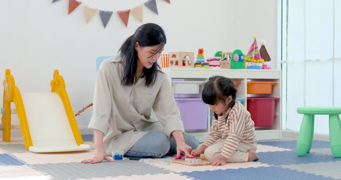 Asian single mother sits on floor with preschool daughter, guiding block play and hand coordination activity in bright playroom, supporting cognitive growth, patience, bonding, early learning at home.