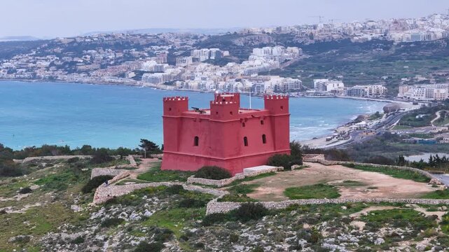 red tower crenellated watchtower hilltop fortification overlooking wetland nature reserve salt marsh pond farmland countryside mellieha rooftops distant malta aerial drone footage rural landscape