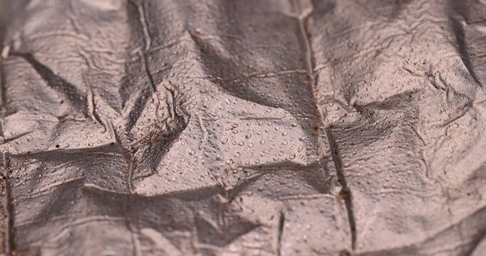 Wet metallic foil texture with fluid bubbles, crumpled aluminum wrap material covered in tiny clear water droplets close up indoors