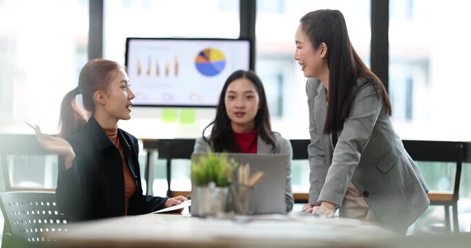 Three young asian businesswomen are collaborating and discussing a project, reviewing financial data charts on a computer screen and documents during a meeting in a contemporary workspace