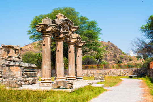 four intricately carved stone pillars at the historic Sahastrabahu Temple in Nagda, Rajasthan, framed by lush green trees, distant hills and a clear blue sky