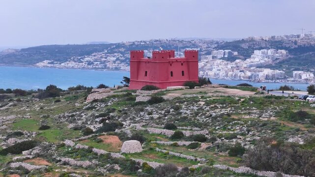 red fortified watchtower headland sweeping sandy bay turquoise water hillside coastal town skyline beachfront promenade parked cars visitors hiking trail coastal road aerial drone footage malta