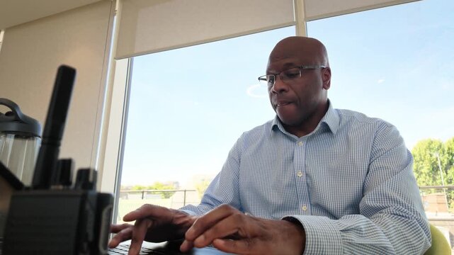 Male office worker in a striped shirt concentrates on a computer task at his desk with daylight pouring through large windows.