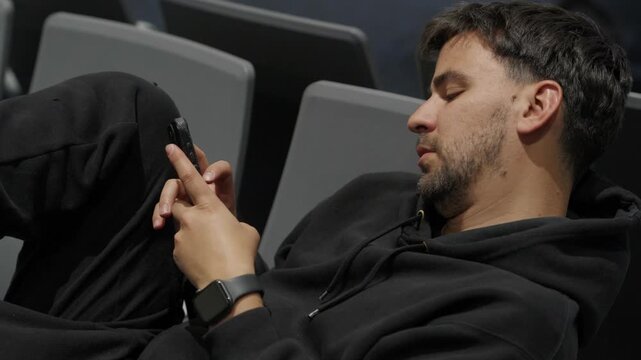 Man in a black hoodie slouched across airport seats scrolling on his phone while waiting