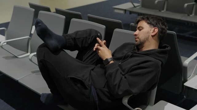 Tired man in black hoodie lying across empty airport gate seats scrolling on his smartphone