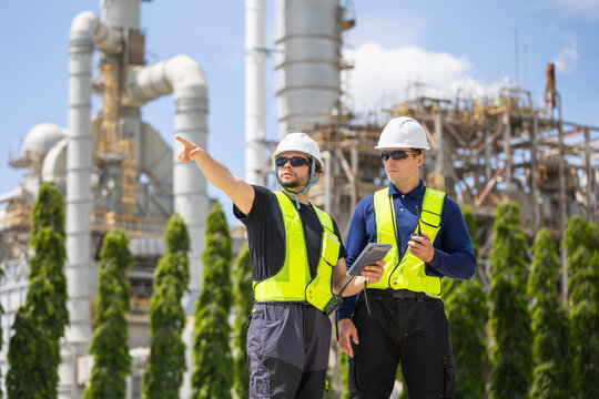 Oil refinery engineers with tablet pointing and discussing at plant, petroleum plant workers in safety gear inspecting industrial facility, energy industry professionals working together outdoors