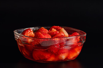 Close-up macro of delicious sliced fresh ripe strawberries with sweet syrup in a transparent square glass bowl against a dark sleek background © Ol-Ni