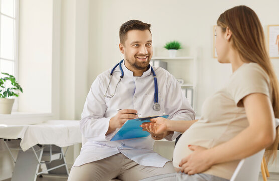 Smiling male doctor in white coat sitting in medical office, holding clipboard and consulting pregnant woman. Happy expectant mother touching belly while discussing health concerns with physician.