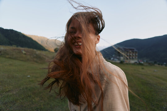 A young woman with brunette hair stands in a windy field, smiling softly as hair sweeps across her face, capturing a candid, carefree lifestyle moment outdoors.