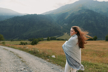 Fototapeta premium Woman standing on a dirt road in a valley wrapped in a light shawl looking to the side with wind in her hair Mountain view outdoor scene travel lifestyle calm mood