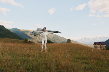 Naklejka premium Man in white clothes stands in a grassy field with a scarf spread wide, enjoying the outdoors, bright day, distant mountains, sense of freedom and adventure