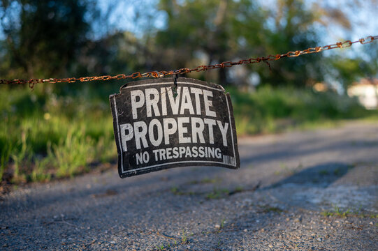 close-up shallow-focus view of a weathered Private Property No Trespassing sign hanging from a strand of barbed wire over a rural asphalt road