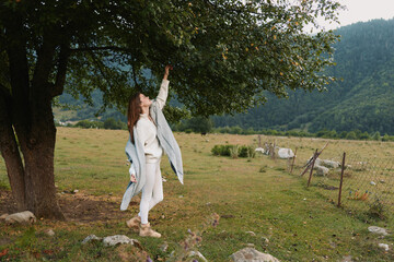 Naklejka premium Woman in a white dress and jacket reaches toward a tree in a rural field, enjoying nature and outdoor life. A peaceful, sunlit moment with grazing cows nearby.