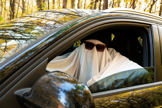 Person wearing ghost costume driving car while holding paper map, searching for direction and navigating route. Sunglasses cap and White fabric disguise covers the body and face, creating humorous