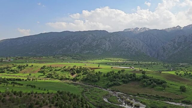 A wide green valley stretches beneath a towering mountain range streaked with snow, its winding stream bordered by scattered trees under a bright cloudy sky
📍 Marivan, Kurdistan, Iran 