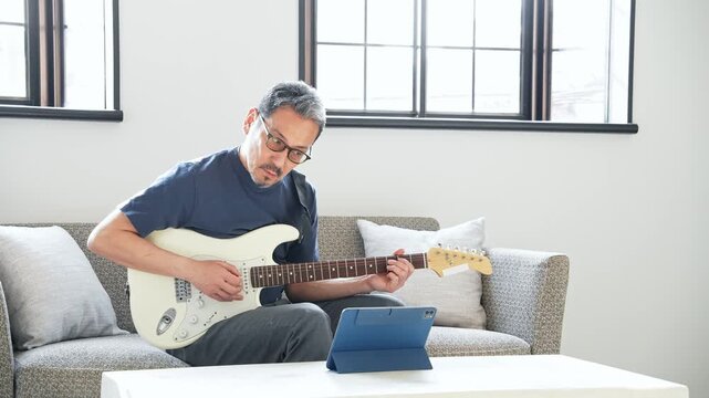 タブレットを見ながらエレキギターを弾く男性。音楽、楽器、演奏、練習、趣味、動画、バンド、音楽家、作曲