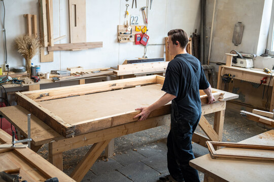 Carpenter preparing wooden window frame for renovation at workbench in carpentry workshop, craftsman disassembling parts of antique window frame with signs of damage at restoring workstation .