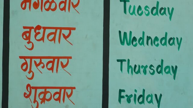 Educational wall chart displays days of the week in Marathi script (orange) and English (green) on light blue background in rural Maharashtra school. Camera tilts downward to reveal weekdays spelling.