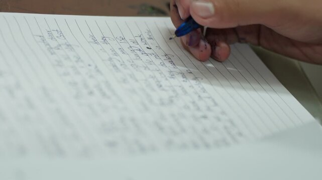 A student's hand holds a blue pen while writing mathematical equations on lined notebook paper during an examination. The close-up shot captures the active writing process with completed calculations.