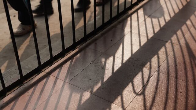 Shadows of children walking are cast through a metal fence onto a paved surface. The ground shows striped shadow patterns from the fence rails as students go to school in Rural India.