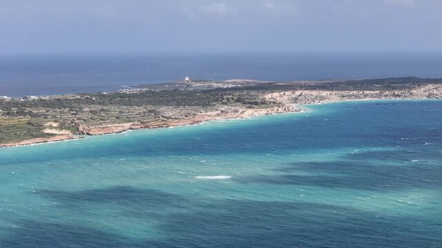 rugged limestone headland malta jagged coastline turquoise shallows fading deep mediterranean blue distant white tower silhouette rocky outcrops aerial drone footage remote peninsula coastal cliffs