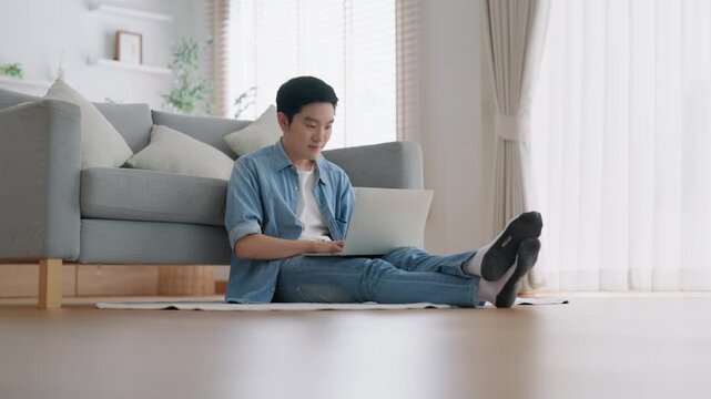 Young Asian man relaxing on comfortable couch, using laptop at home, happy boy chatting with friends in social network, spending weekend, watching movie, shopping online