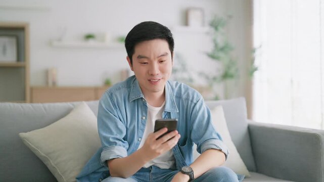 Smiling handsome young Asian man sitting on cozy couch, using funny mobile apps in living room. man at home, doing online shopping, messaging friends, posting on social media