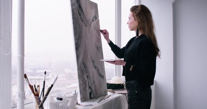 Acrylic paint. Artist working. Talented woman master using palette knife tool for abstract canvas near window in studio.