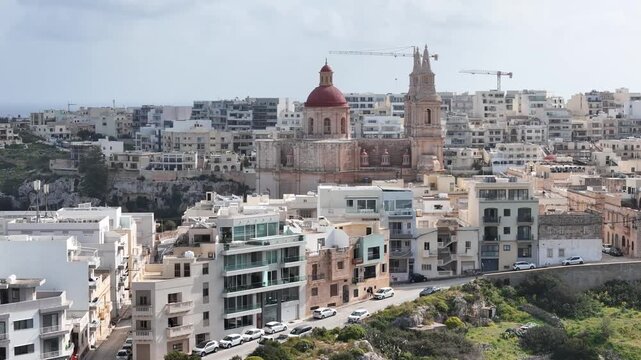 parish church red dome mellieha bell towers baroque facade construction cranes background rooftops apartment buildings coastal skyline mediterranean sea malta aerial drone footage close orbit shot