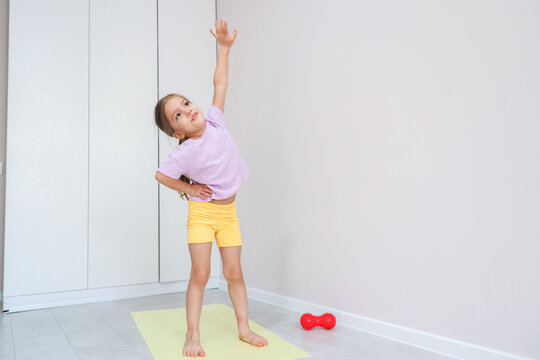 Child Practices Balance And Flexibility On Pale Mat In Clean Modern Room, Standing Barefoot Beside Exercise Ball While Reaching Upward, Showing Healthy Active Lifestyle, Coordination