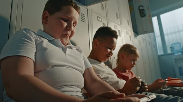 Happy Primary School Classmates Sitting on Floor Beside Personal Lockers with Handheld Electronics During Break. Authentic School Atmosphere, Teamwork, Communication, and Everyday Student Life.
