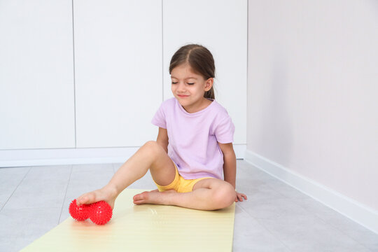 Small Girl In Pastel Clothes Sits On Mat Indoors While Pressing Foot On Red Spiky Massage Ball, Showing Gentle Sensory Exercise, Relaxation, And Healthy Home Fitness
