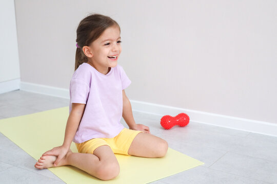 Cheerful Young Girl Sits On Yoga Mat In Bright Minimalist Room, Smiling During Gentle Stretching Practice Beside Red Massage Roller, Creating Playful Wellness Scene For Home Exercise