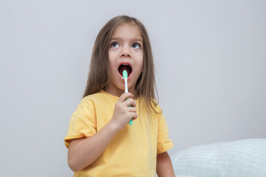 Young Girl Brushing Teeth With Toothbrush Indoors Against Light Gray Wall, Wearing Yellow Shirt And Looking Upward While Keeping Mouth Open. Additional Visual Context For, With Clear Visible Detail