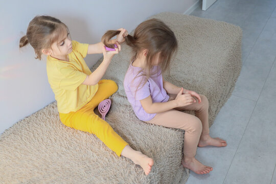 Two young girls sitting on soft rug while one styles other child hair in bright home interior, casual playtime grooming moment with pastel clothes and gentle domestic scene