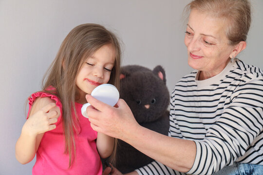 Older Woman Smiles While Helping Young Girl Hold Compact Mirror During Quiet Beauty Play At Home, With Soft Natural Light, Striped Shirt, Pink Top, Plush Toy Background, Warm Family