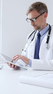 Middle aged doctor man wearing lab coat and stethoscope, is using digital tablet while sitting at desk in medical office, with medical chart and pen in the foreground. Medicine concept