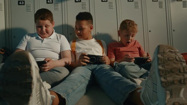 Elementary School Children Sitting in Sunny Corridor Beside Metal Lockers while Using Portable Gaming or Learning Devices. Modern Education, Digital Lifestyle, Childhood Communication. Handheld.