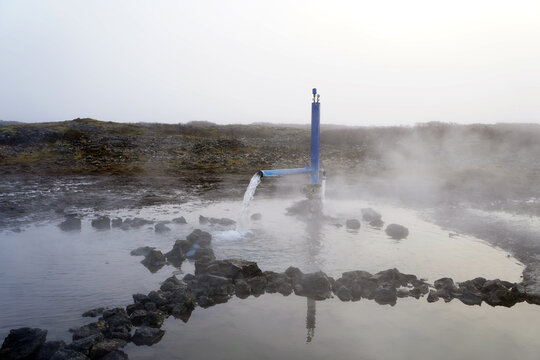 A pipe pours hot geothermal water into a stone-lined thermal pool, sending steam into the cool air across a barren volcanic landscape in Iceland